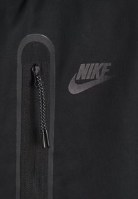 Svart Nike-väska med texturerat tyg, vertikal dragkedja med paracord-dragflik och reflekterande logodetalj.