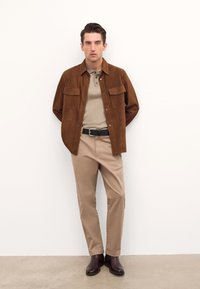 Ruskosuinen mokkajakku päällä beige polo, yhdistettyinä beigeihin housuihin ja ruskeisiin nahkasaappaisiin. Yksinkertainen muotoilu ja neutraali väriasteikko.