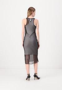 Schwarzes, transparentes, ärmelloses Kleid mit figurbetonendem Design, Schlüsselhenkel-Rückendetail und knielanger Länge. Dazu getragen mit Riemchen-Pumps.