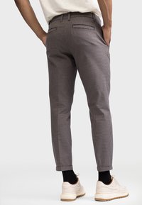 Hnědé slim-fit kalhoty s kostkovaným vzorem, vyrobené z lehkého materiálu. Mají dvě zadní kapsy a tapered design nohavic. Kombinované s bílými teniskami.
