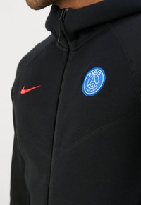 Sort zip-up hoodie med et hævet Paris Saint-Germain logo i blå og en rød Nike swoosh. Glat tekstur med atletiske fit sømme.