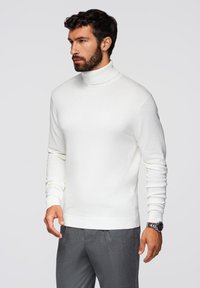 Biała, dopasowana sweter w serek z miękkiego dzianiny, z długimi rękawami i ściągaczami na mankietach, w połączeniu z szarymi spodniami i zegarkiem.