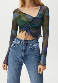 Top trasparente a maniche lunghe nei toni del verde e blu, con un design arricciato e lacci regolabili in vita, abbinato a jeans in denim a vita alta.