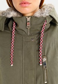 Parka verde oliva con un ribete de capucha de piel sintética, presenta cremalleras dobles, cordones con un patrón rojo, negro y blanco, y acentos metálicos.