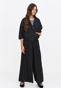 Czarna krótka kurtka z kołnierzem stójkowym, z zamkiem z przodu i kieszenią boczną; noszona na czarnych szerokich spodniach; model w płaskich butach.