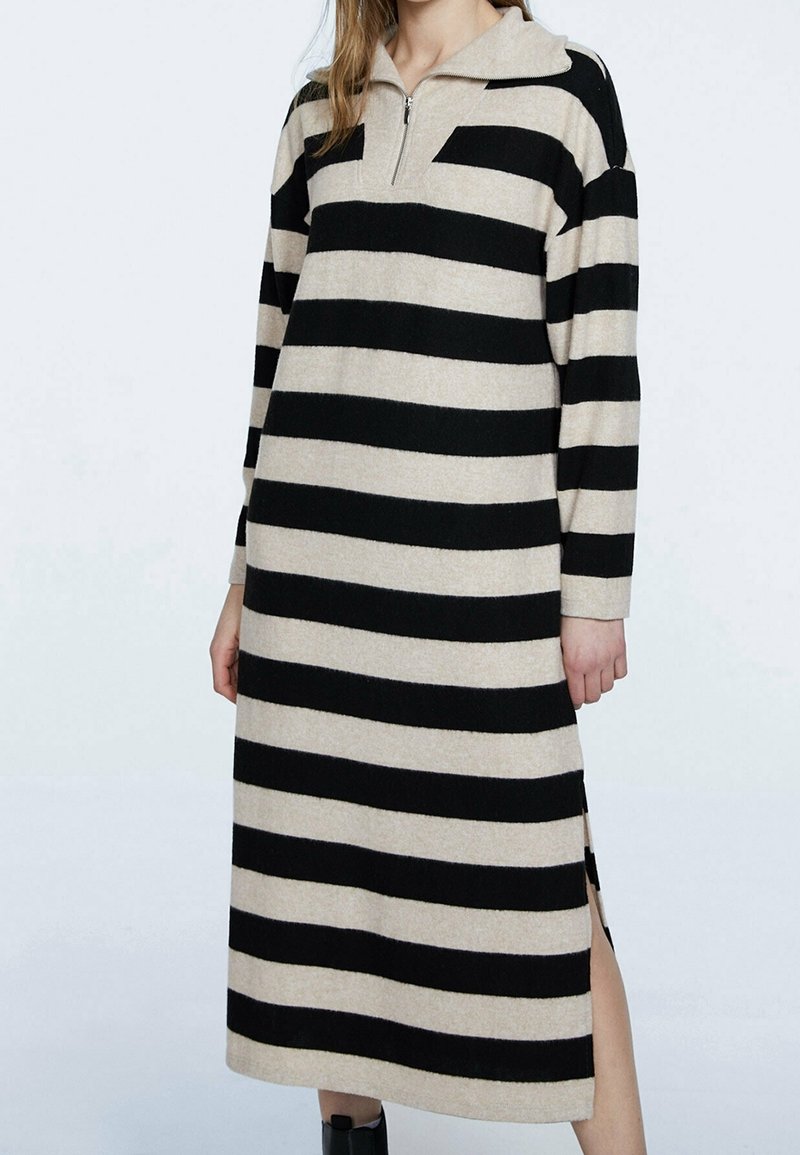 Abito lavorato a maglia a righe con una combinazione di colori nero e beige, maniche lunghe, colletto con zip e spacchi laterali per facilitare i movimenti.