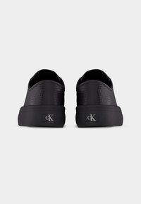 Baskets basses noires avec motif texturé, semelles épaisses et logo "cK" sur le talon, vues de dos sur un fond uni.