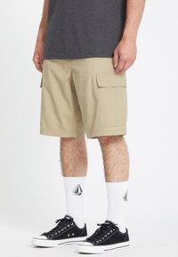 Pantalones cortos de cargo beige con bolsillos laterales, llevados con zapatillas negras y calcetines blancos con un logo. Camiseta gris parcialmente visible.