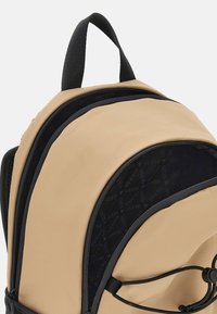 Beige och svart ryggsäck med en slät yta, som har en rundad design, flera dragkedjefack och logotypdetaljer på tyget.