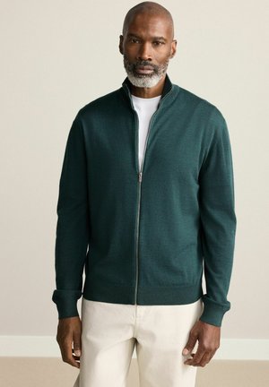 Dunkelgrüner Reißverschluss-Cardigan aus weichem Stoff, mit hohem Kragen und gerippten Bündchen. Über einem weißen T-Shirt und hellbeigen Hosen getragen.