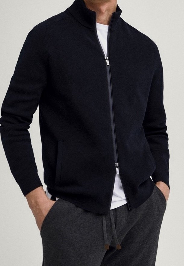 Homme portant une veste zippée noire sur une chemise blanche et un pantalon gris foncé à cordon, les mains dans les poches, debout devant un fond clair.