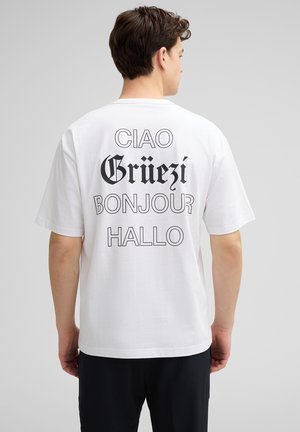 Witte katoenen T-shirt met grote tekst op de achterkant: "Ciao," "Grüezi," "Bonjour," en "Hallo" in wisselende lettertypes en groottes.