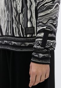 Pull en tricot noir et blanc avec des motifs de vagues abstraites, un tissu texturé et des poignets côtelés avec une étiquette de marque en tissu.