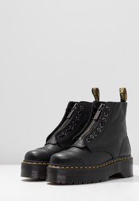 Bottes à plateforme en cuir noir avec des zips à l'avant, des coutures jaunes et des onglets de traction avec logo sur une surface et un fond blanc uni.