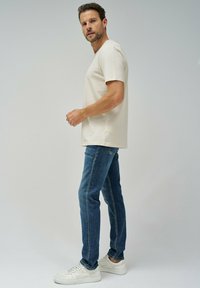 Camiseta color beige de cuello redondo, jeans azul ajustados y zapatillas blancas. La camiseta tiene mangas cortas; los jeans tienen un corte relajado y un sutil pespunte.