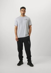 Šedé tričko s krátkým rukávem a grafickým potiskem na hrudi, doplněné o černé tepláky s logem na boku a černé tenisky.