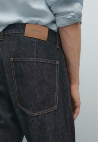 Nærbillede af en persons ryg, iført mørke denimjeans med orange syninger. En tan læderlabel er synlig på taljebåndet.