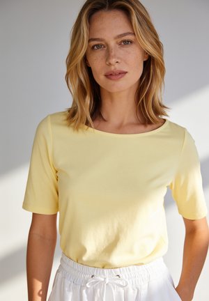 Junge Frau mit schulterlangem blonden Haar, die ein gelbes kurzärmeliges Shirt und weiße Hosen mit Kordelzug trägt, steht vor einem hellen Hintergrund.