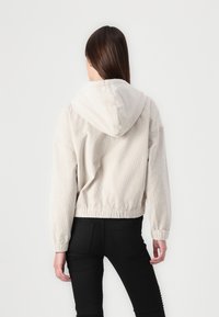 Kvinna med långt brunt hår, klädd i en ljusbeige kordjacka med huva och svarta byxor, som står vänd bort från kameran mot en vit bakgrund.