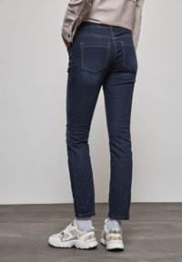 Mörkblå denimjeans med smal passform, med två bakfickor och diskreta sömnadsdetaljer. Bärs med vit- och grå sneakers.