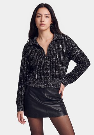 Vrouw met lang donker haar, gekleed in een zwart-wit gebreid vest en een zwarte leren minirok tegen een effen achtergrond.