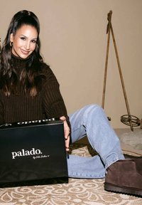 Zwarte rechthoekige doos met "palado" in witte tekst. Bruine trui, lichtblauwe spijkerbroek en bruine suède laarzen. Geprinte vloerkleed en neutrale achtergrond.