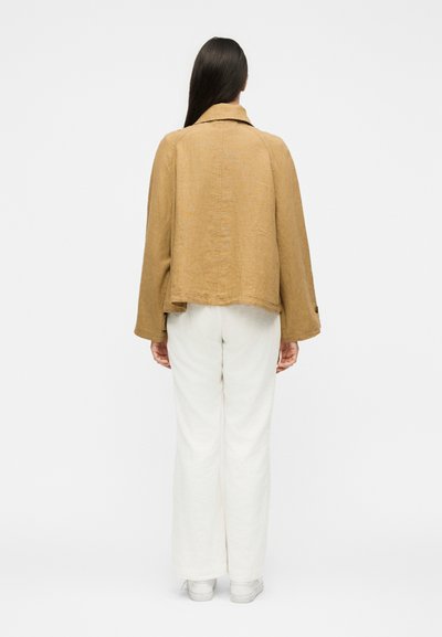 Γυναίκα με μακριά σκούρα μαλλιά που φοράει φαρδιά μπεζ ζακέτα, άσπρο παντελόνι και άσπρα παπούτσια, στέκεται γυρισμένη με την πλάτη προς ένα απλό φόντο.