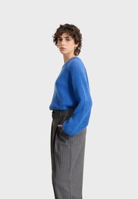 Blauer, flauschiger Pullover mit lockerem Schnitt und weiten Ärmeln, kombiniert mit grauen Hose mit Streifen, hervorgehoben durch eine hohe Taille und einen Gürtel.
