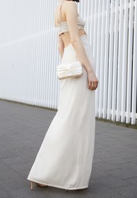 Beżowa sukienka maxi z wycięciami, dobrana z kremową kopertówką z węzłowym detalem oraz beżowymi wysokimi obcasami. Materiał o fakturze.