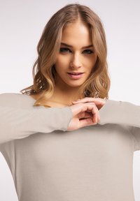 Lichtbeige langarmtop met een ronde hals, zachte stofstructuur en licht verweerde mouwranden, gedragen door een model.