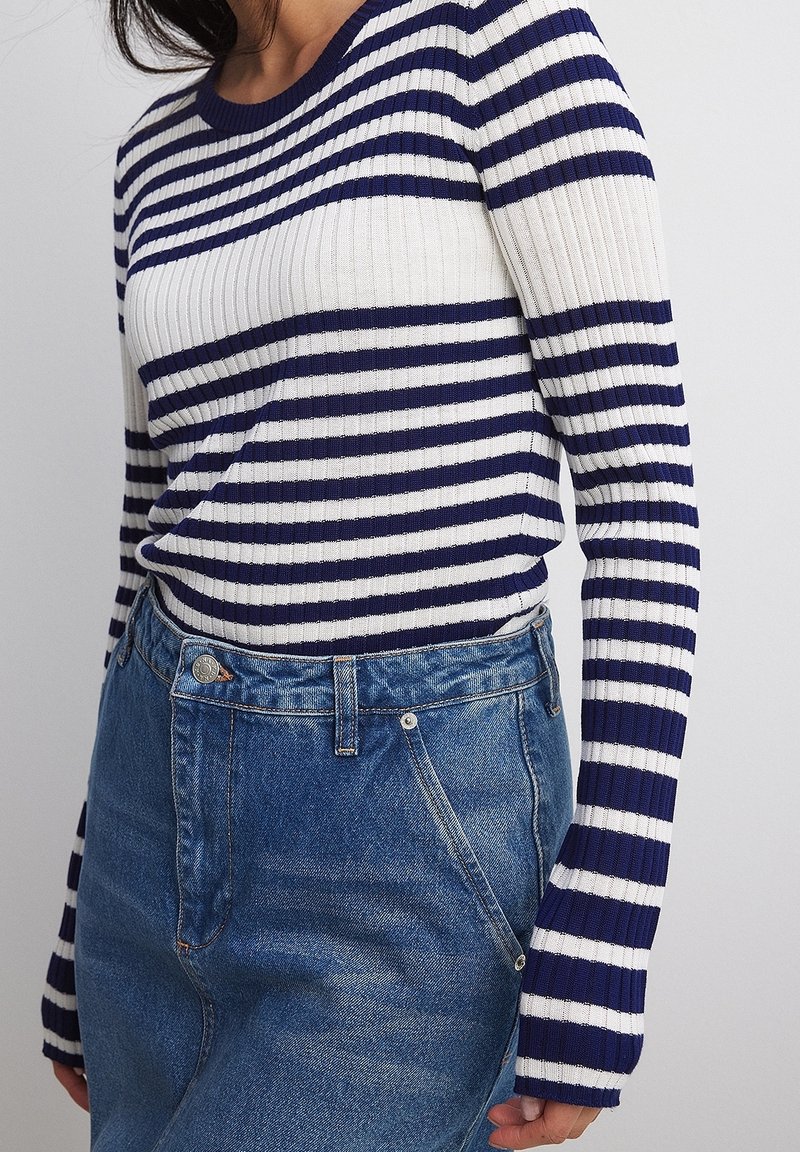 Femme portant un haut côtelé à manches longues rayé bleu marine et blanc, rentré dans un jean en denim bleu taille haute, sur un fond uni.