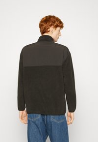 Mørkegrønn fleecejakke med høy krage, lange ermer og glatt tekstur, kombinert med blå denimjeans.