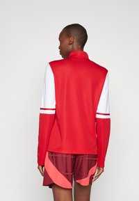 Rød og hvid langærmet atletisk top med høj krave, der har stribede detaljer på ærmerne, parret med korte bordeaux shorts.