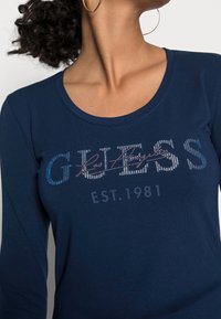 Granatowa koszulka z długim rękawem z mieszanki bawełny; posiada świecące logo "GUESS" oraz nadrukowane w kontrastowym kolorze "EST. 1981" poniżej.