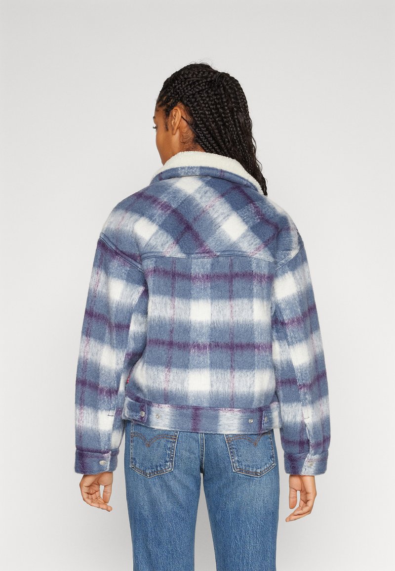 Veste à carreaux présentant des teintes bleu, blanc et violet. Tissu doux avec un col en fausse fourrure et des boutons-pression, associée à un jean en denim.