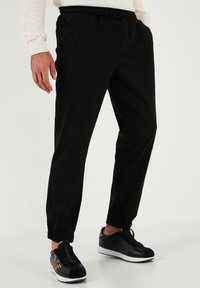 Schwarze Jogginghosen mit elastischem Bund und krempelbaren Knöcheln, aus leichtem Stoff gefertigt. Kombiniert mit schwarzen Sneakers mit Designelementen.