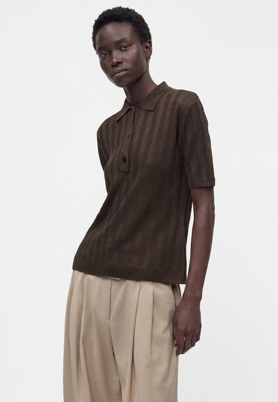 Femme portant un polo marron à manches courtes avec des rayures verticales et un pantalon plissé beige, debout devant un fond blanc uni.