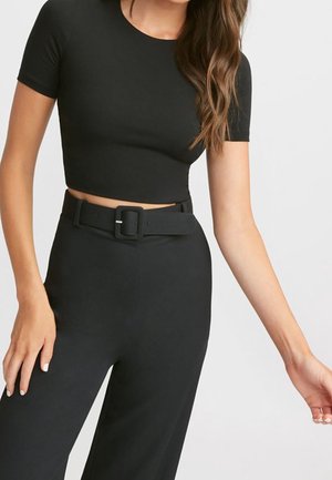 Kobieta ubrana w dopasowany czarny krótki top i czarne spodnie z wysokim stanem, szerokim czarnym paskiem i prostokątną klamrą.