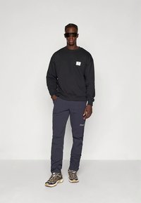 Czarna bluza z naszywką z logo, granatowe spodnie z detalem logo oraz masywne beżowo-czarne sneakersy. Modelka w okularach przeciwsłonecznych.