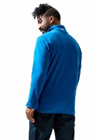 Blauwe fleece pullover met een hoge kraag, textuurstof en een klein logo bij de nek, gecombineerd met donkere broeken en een relaxte pasvorm.