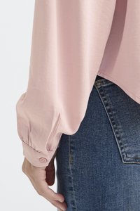 Blusa lunga a maniche con polsini arricciati di un delicato rosa, abbinata a jeans in denim blu scuro. La blusa presenta una vestibilità comoda e una texture morbida.