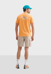Camiseta de algodón naranja con estampado en la espalda, combinada con pantalones cortos cargo beige con un patrón de hojas. El modelo lleva una gorra azul y zapatillas multicolores.