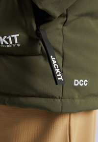 Podszewka oliwkowa kurtka z teksturowanej tkaniny, z czarnym uchwytem zamka błyskawicznego, białym logo oraz kontrastującą tanową dolną częścią.