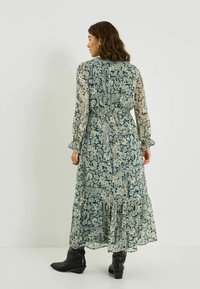 Robe longue et fluide à imprimé floral bleu marine et crème, avec des manches longues transparentes et un ourlet à volants. Portée avec des bottines noires à la cheville.
