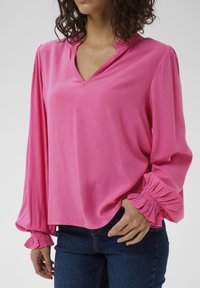 Blouse rose avec un col en V et des poignets froncés. Fabriquée en tissu lisse, elle présente des manches longues et une coupe décontractée. Associée à un denim bleu.