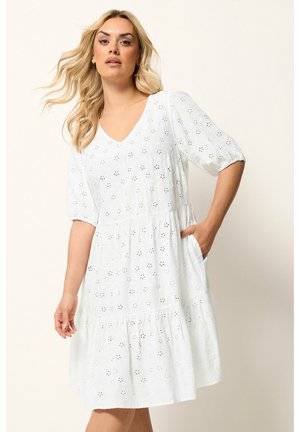 Blondynka ubrana w białą, sięgającą do kolan sukienkę z ażurowym wzorem, krótkimi bufkami, dekoltem w serek oraz kieszenią po boku, stojąca na jednolitym tle.