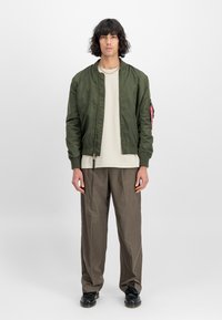 Casaco bomber verde com fecho à frente, punhos canelados e um patch na manga, combinado com uma t-shirt de cor clara e calças castanhas.
