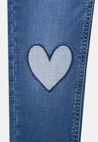 Jeans in denim con una toppa a forma di cuore blu con cuciture a contrasto. Il tessuto ha una finitura stonewash classica e una texture liscia.