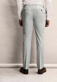 Pantalones ajustados de color gris claro con un frente plano, dos bolsillos traseros con botones y un acabado limpio en los dobladillos. Llevados con zapatos marrones.