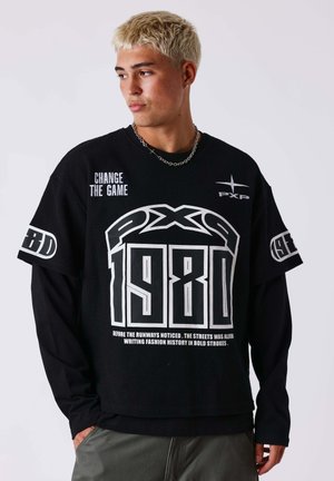 Joven con cabello rubio corto que lleva una camisa negra con capas con texto "PXP 1980" y "Change the Game", mirando hacia un lado.